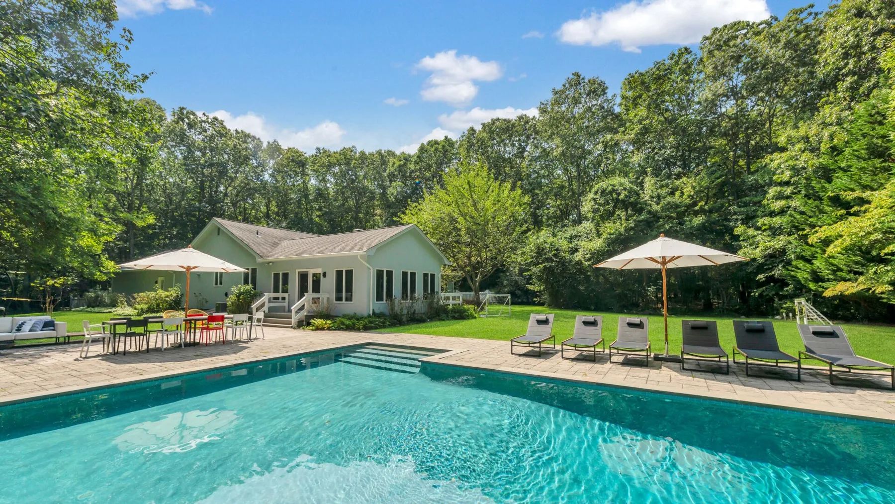 An outdoor swimming pool sits beside a house, with lounge seating and umbrellas.