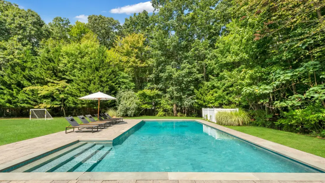 An outdoor swimming pool is bordered by a paved deck, lounge seating, and trees.