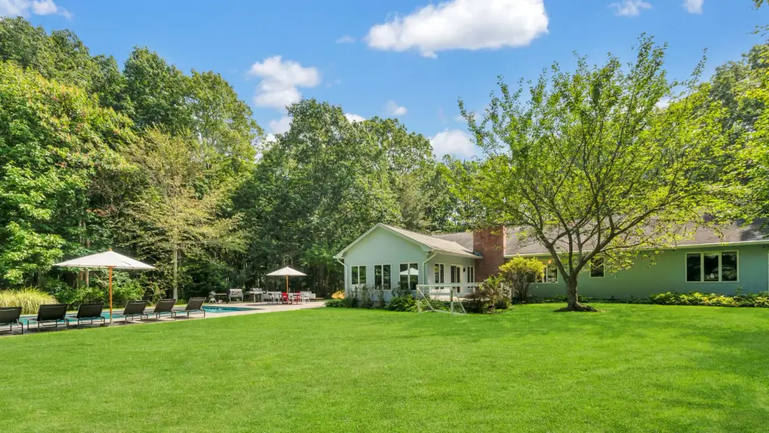 A low house sits beside a broad green lawn, with leafy trees behind it.