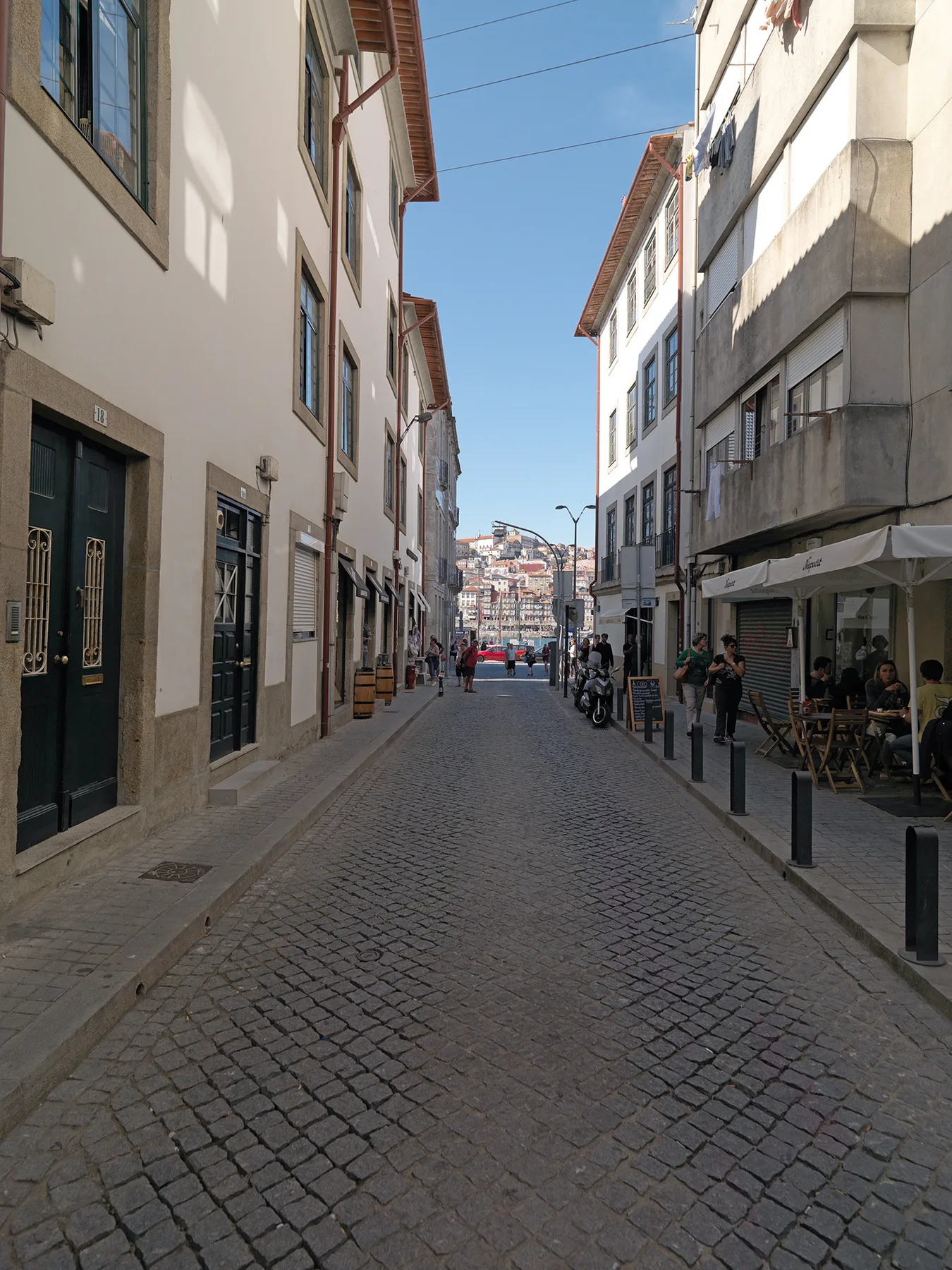 A cobblestone street runs between tall buildings, with a café seating area along the sidewalk.
