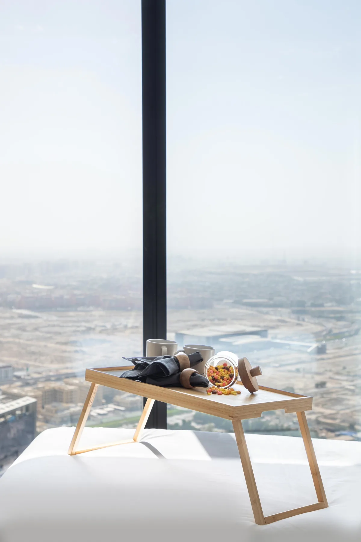 Una bandeja de desayuno está sobre una cama junto a una gran ventana con vistas a la ciudad.