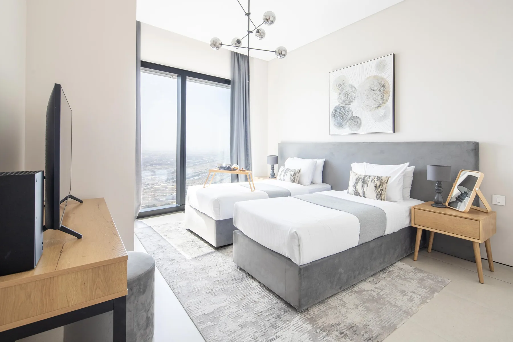 Un dormitorio luminoso con dos camas, grandes ventanas y un cabecero gris tapizado.