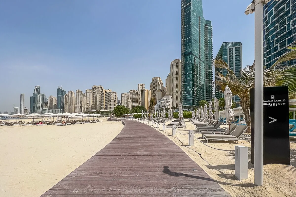 Una pasarela de madera cruza una playa de arena junto a tumbonas y un horizonte urbano.