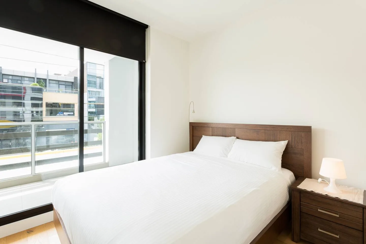 Un dormitorio con una cama junto a una ventana amplia con vista a edificios cercanos.