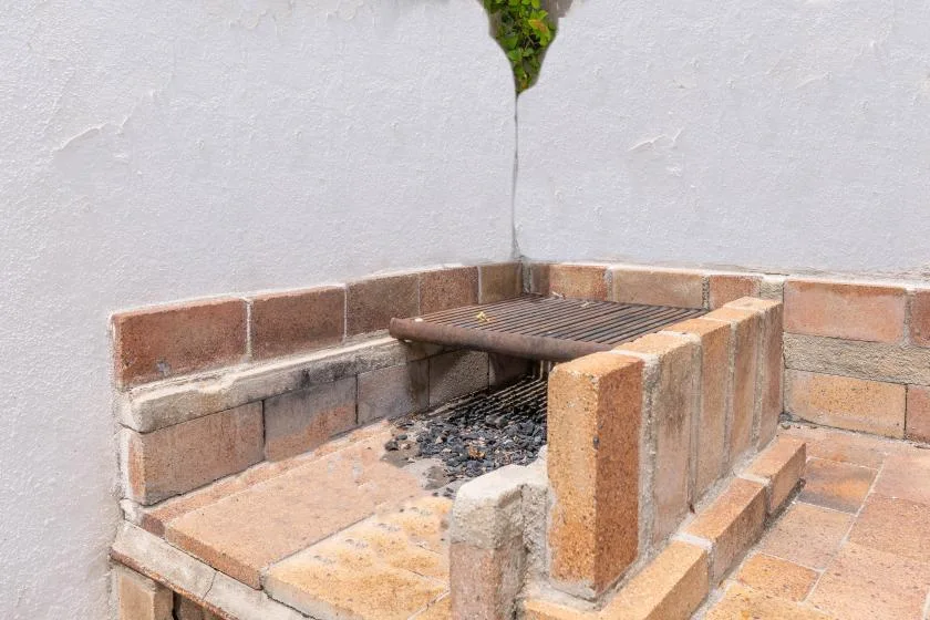 An outdoor brick barbecue with a metal grill sits against a white wall.