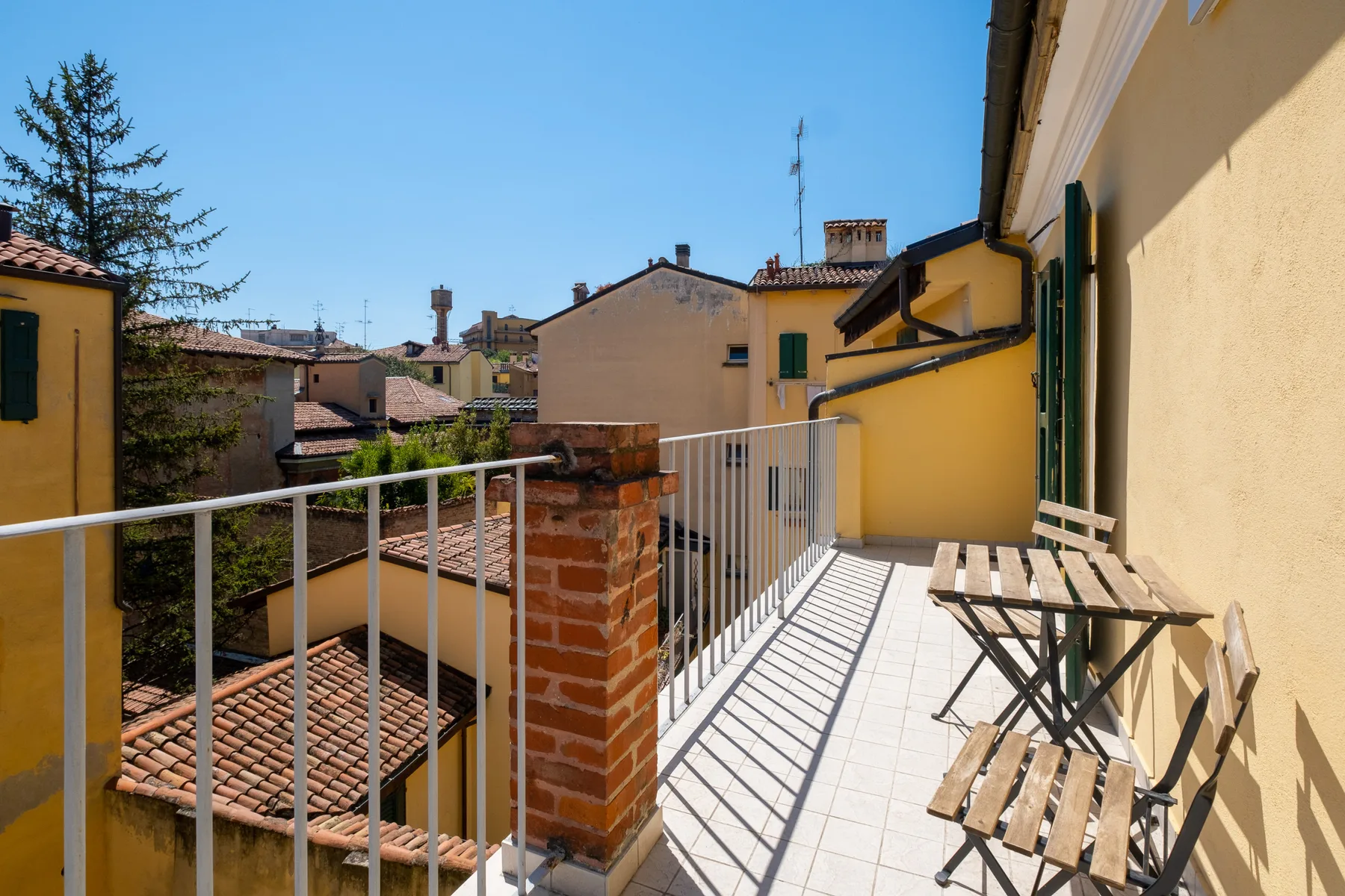 A sunny tiled balcony with metal railings and wooden seating overlooks nearby rooftops.