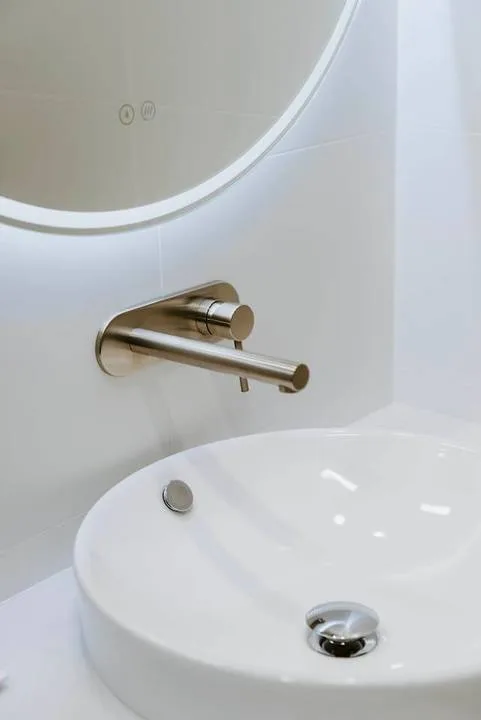 Un lavabo blanco de baño está bajo un espejo redondo, con un grifo de pared.