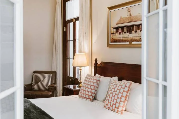 Un dormitorio con una cama tendida, una lámpara de noche y un sillón junto a la ventana.