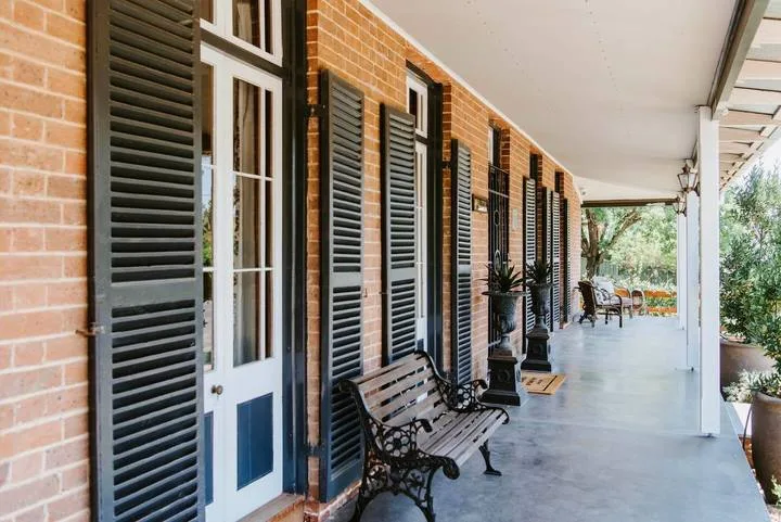 Una galería cubierta recorre un edificio de ladrillo con puertas y ventanas con contraventanas.