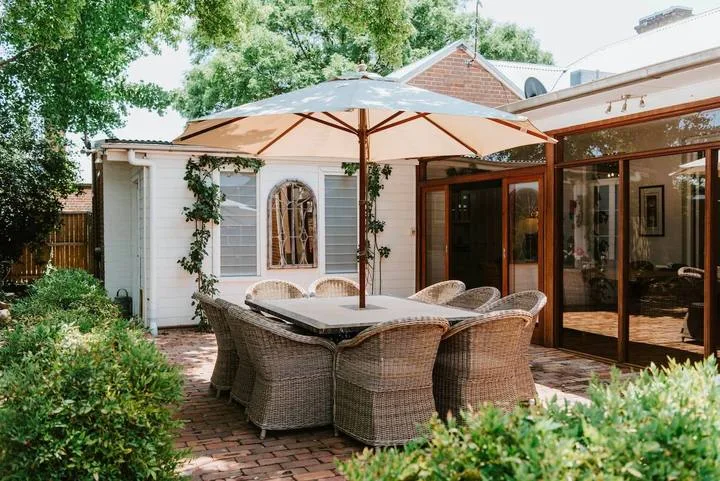 Una zona de comedor en un patio de ladrillo está bajo una gran sombrilla junto a la casa.