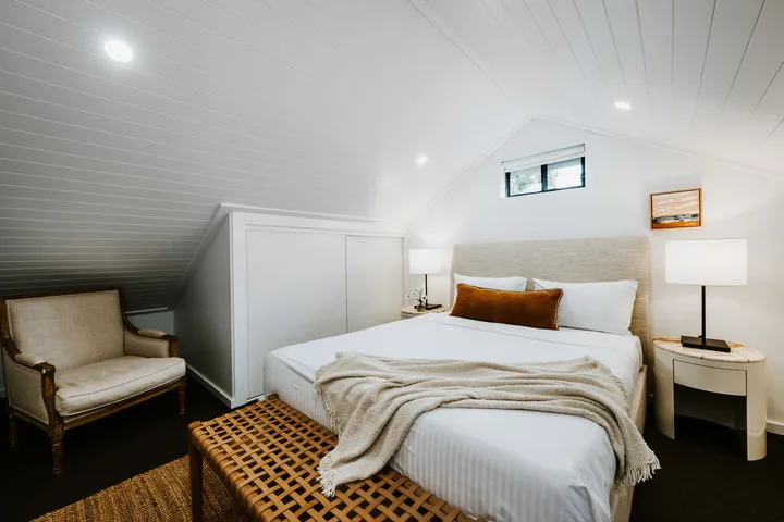 Un dormitorio con cama, mesitas de noche y un sillón bajo un techo inclinado.
