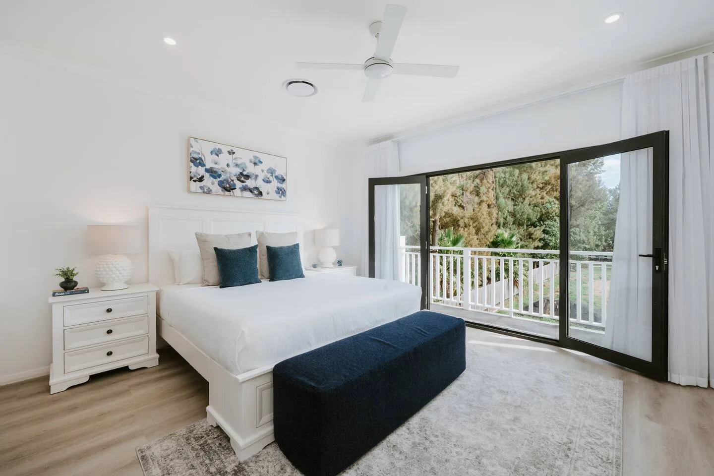Dormitorio luminoso con cama, banco tapizado oscuro y puertas correderas de vidrio que dan a un balcón.