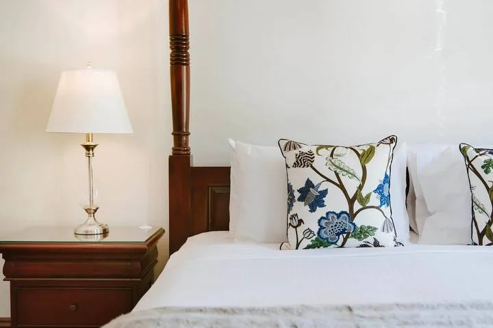 Un dormitorio con ropa de cama blanca, almohadas florales y una lámpara de noche.