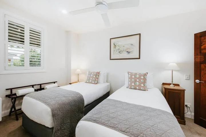 Un dormitorio luminoso con dos camas, mesitas de noche y una ventana con persianas.