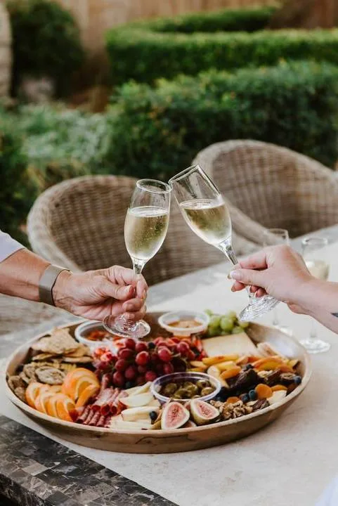 Unas manos alzan copas de vino espumoso sobre una bandeja de frutas y aperitivos en una mesa exterior.