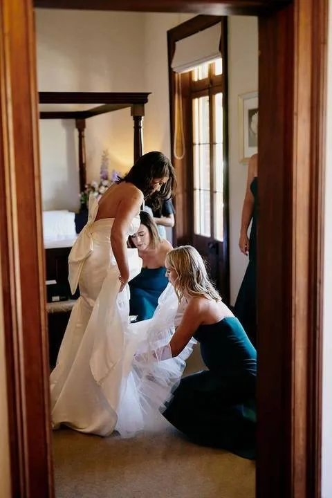 Varias personas ayudan a una novia a acomodar su vestido en un dormitorio.