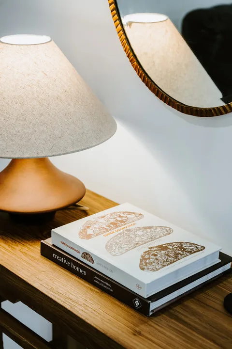 Una mesa auxiliar sostiene una lámpara y varios libros bajo un espejo de pared.