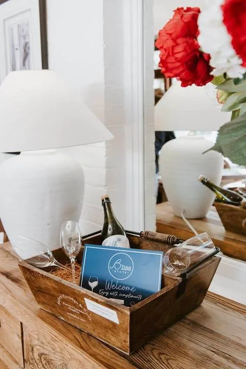 Una bandeja de madera con una tarjeta de bienvenida y una botella está junto a una lámpara blanca y flores rojas.