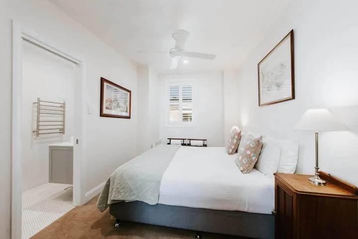 Un dormitorio luminoso con una cama tendida y cuadros en la pared.