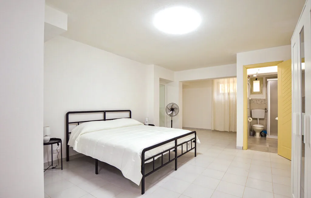 Un dormitorio luminoso con una cama, un ventilador de pie y una puerta abierta al baño.