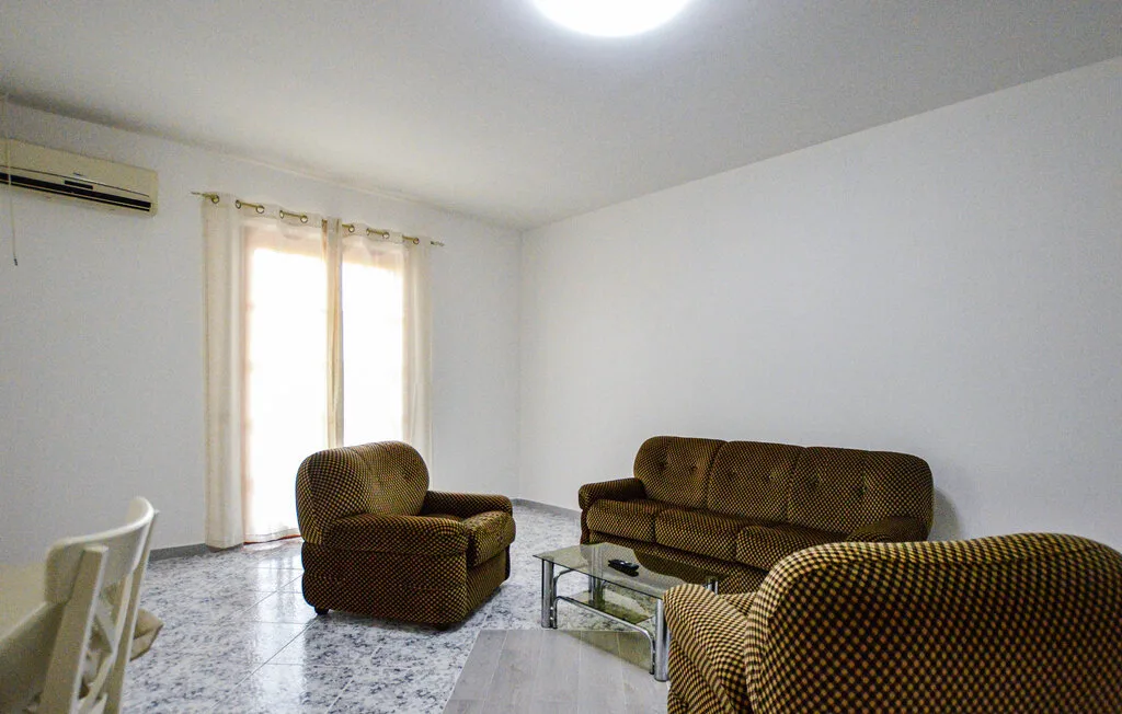 Una sala de estar con asientos marrones, una mesa de centro de vidrio y una ventana luminosa con cortinas.