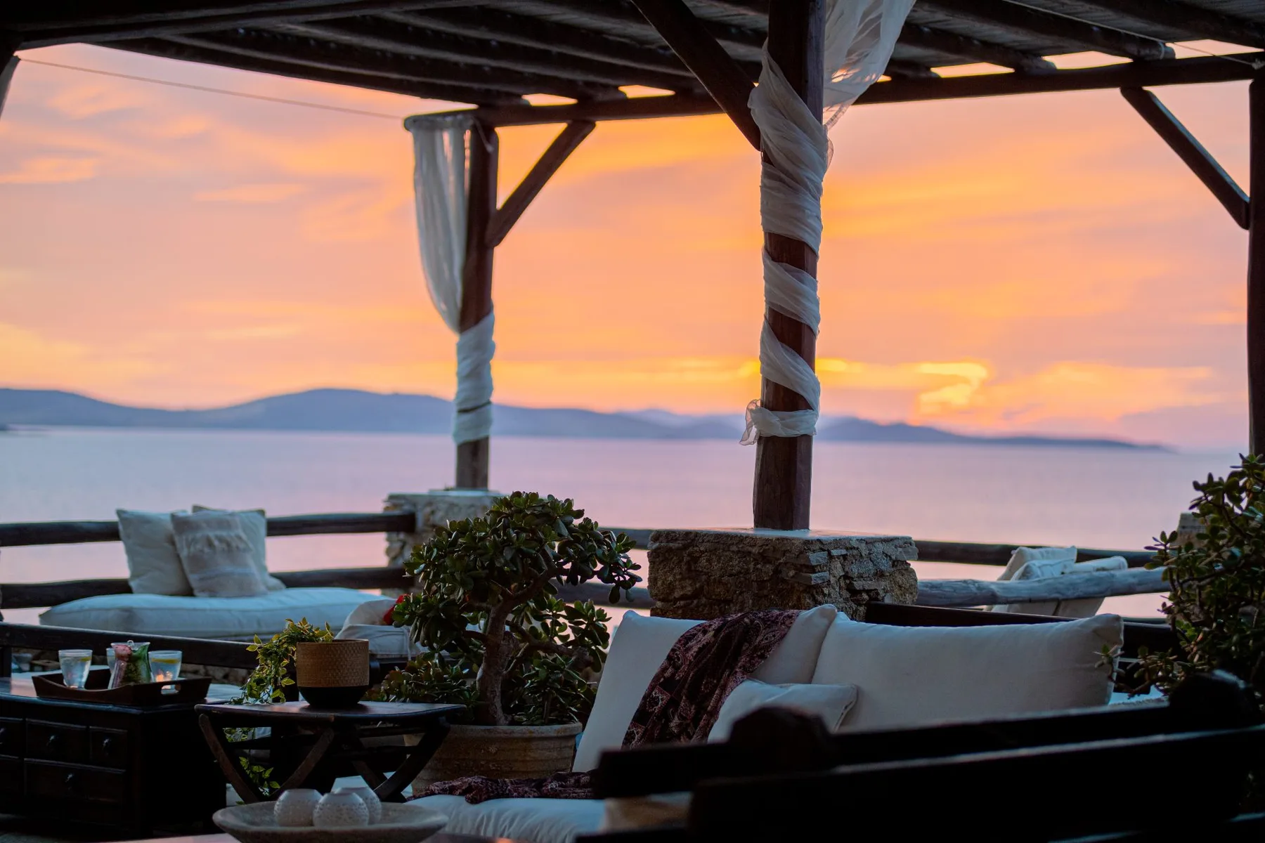 An outdoor lounge overlooks the water at sunset beneath a wooden pergola.