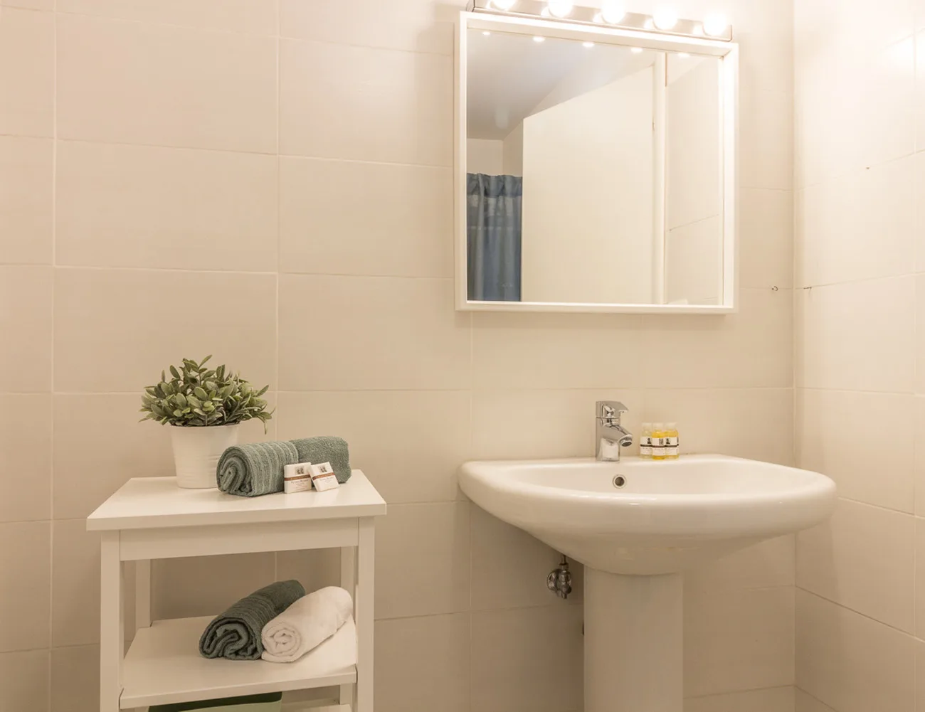 Un baño alicatado con lavabo, espejo y una pequeña mesa con estantes.