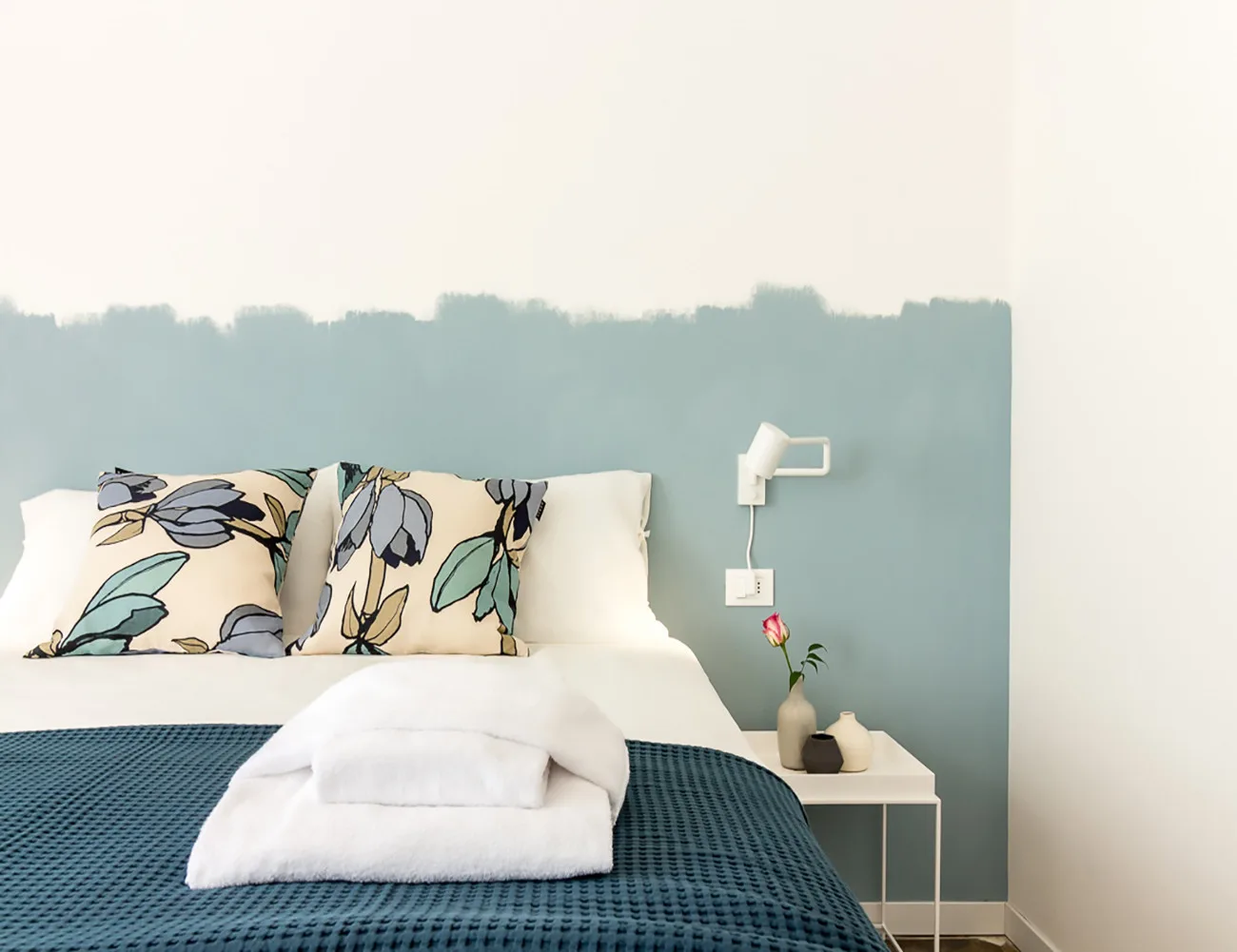 Un dormitorio con almohadas florales y ropa de cama blanca frente a una pared azul apagada.