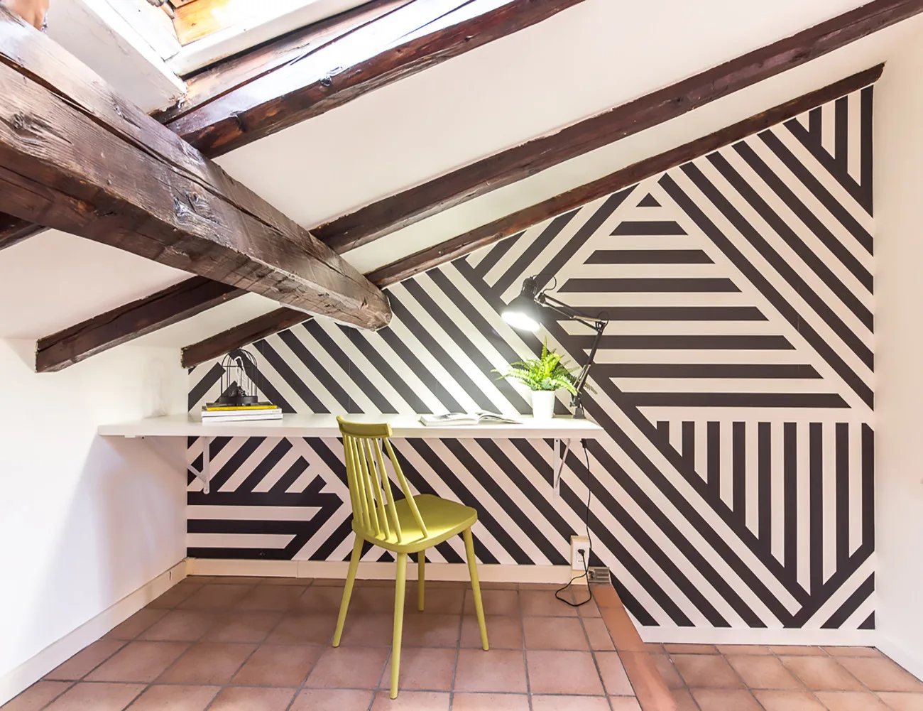 Un pequeño escritorio está frente a una pared geométrica, con una silla verde y una lámpara.