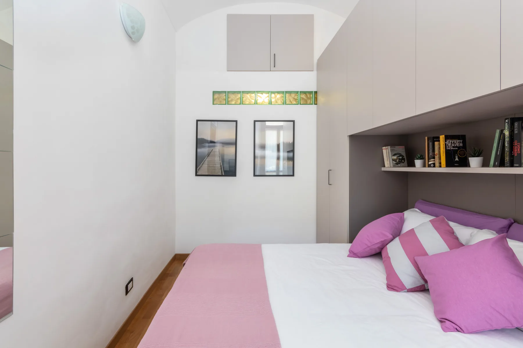 A bedroom with a bed dressed in white and pink bedding beneath built-in shelving.