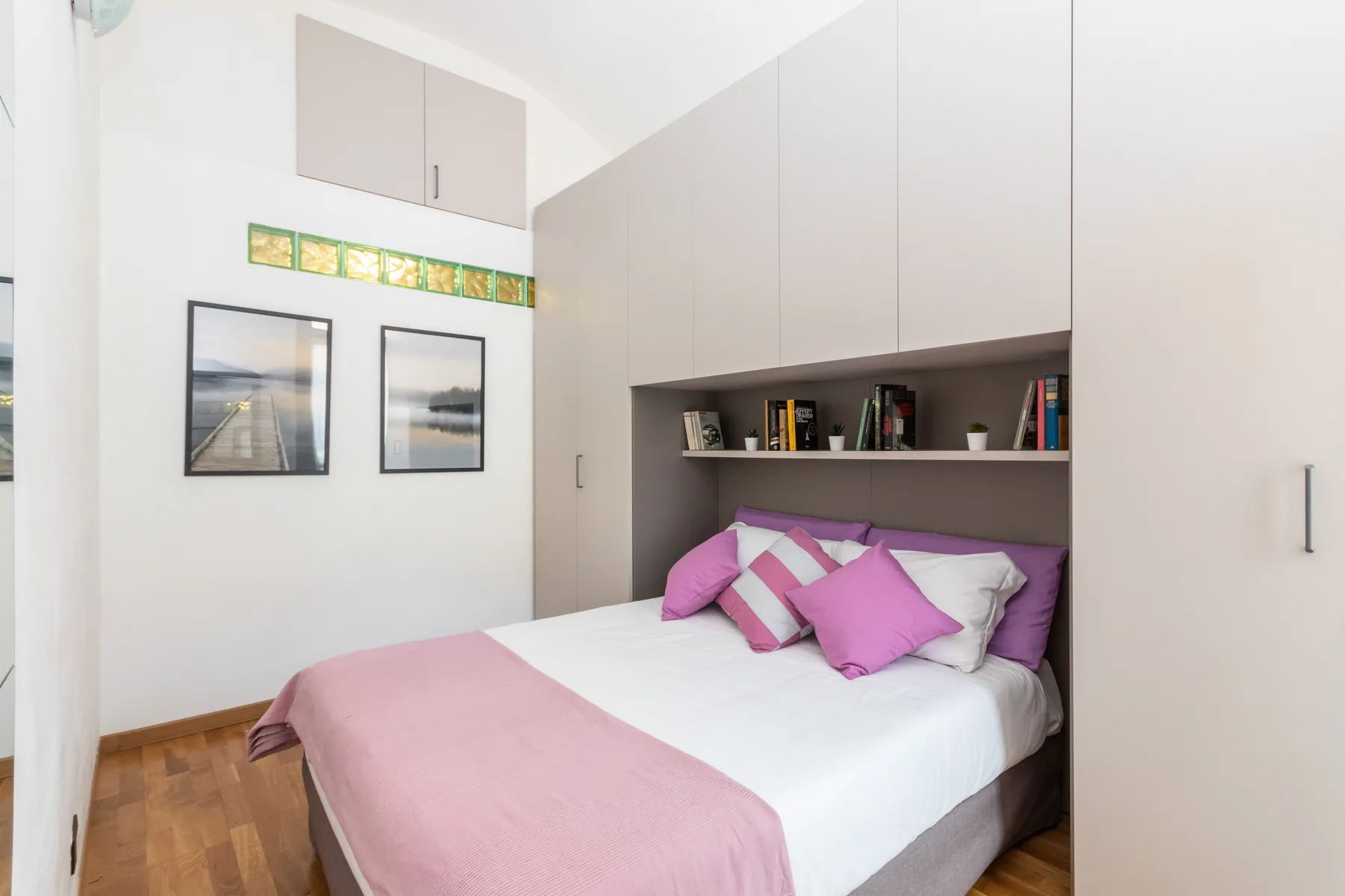 A bedroom with a bed dressed in white and pink beneath built-in shelving.