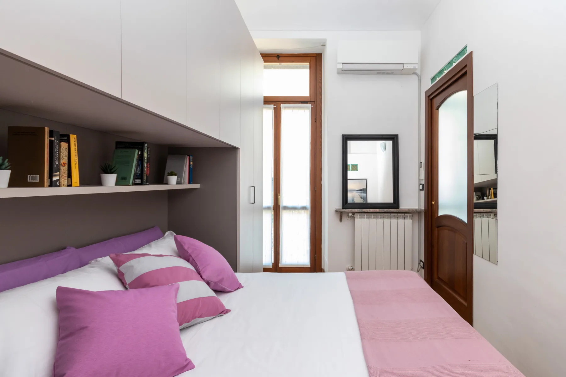 A bedroom with a bed beneath built-in shelving and a tall window.