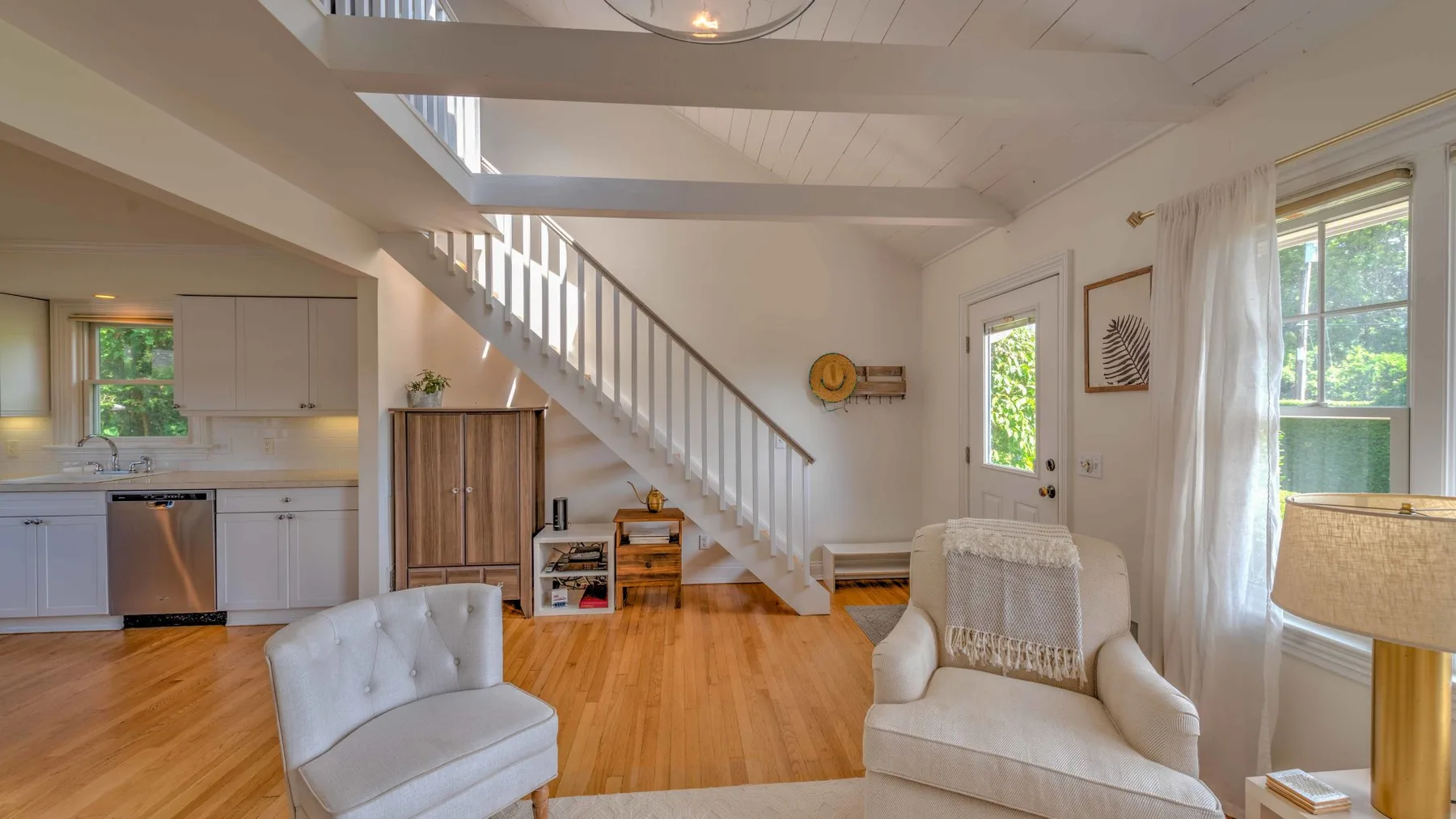 A bright living room with a staircase, light seating, and an open kitchen.