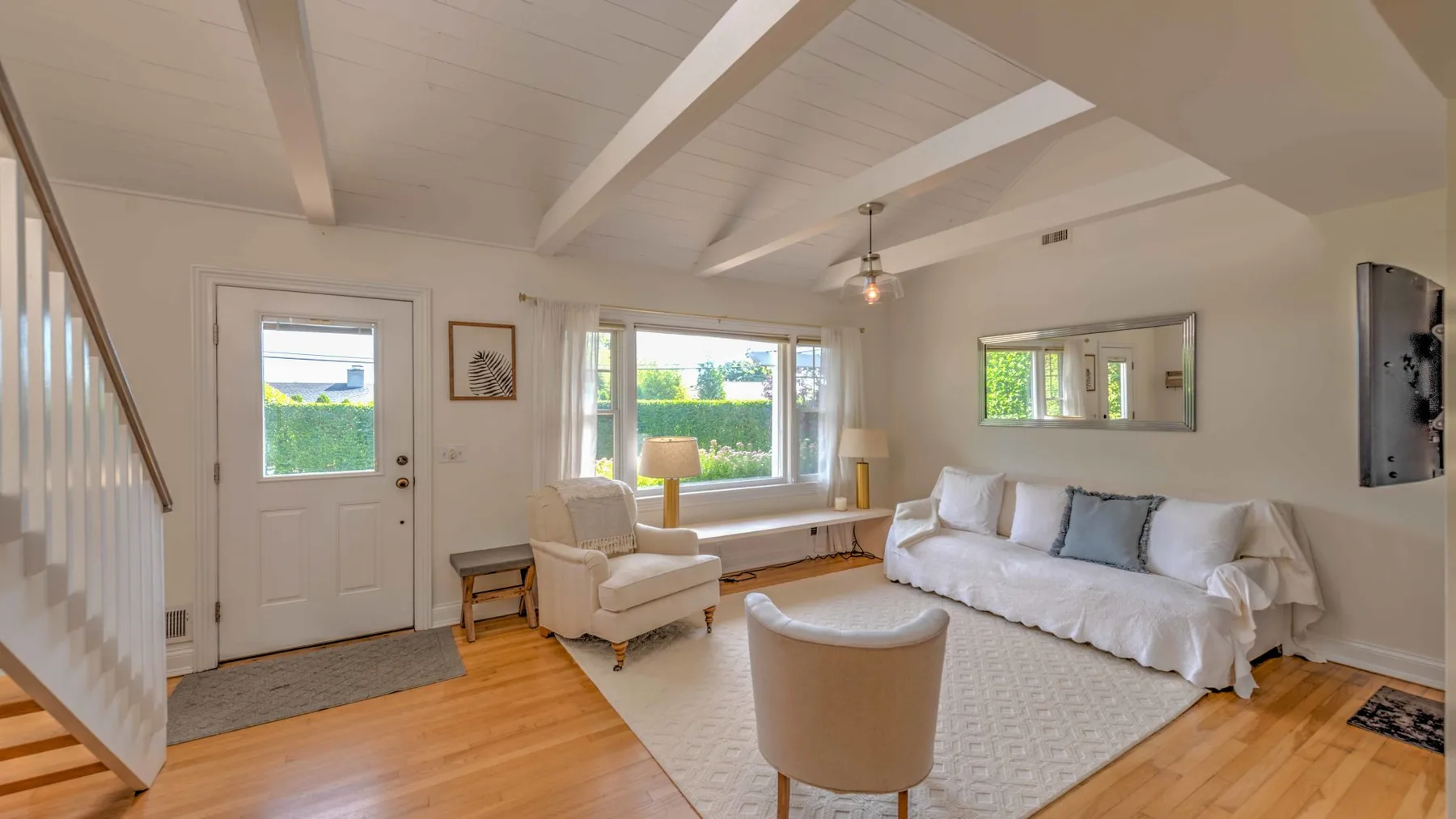 A bright living room with white seating, a large window, and exposed ceiling beams.