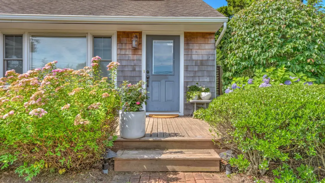 A blue front door opens onto a small wooden porch surrounded by flowering shrubs.