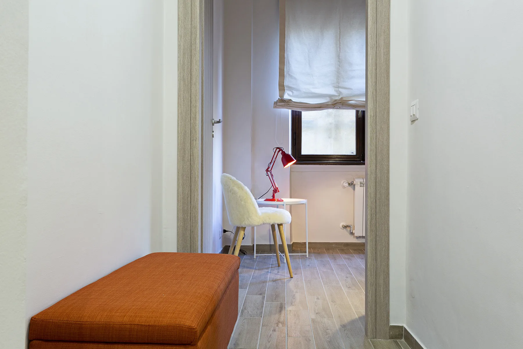 A bright room with a desk, chair, and red lamp sits beyond a hallway.