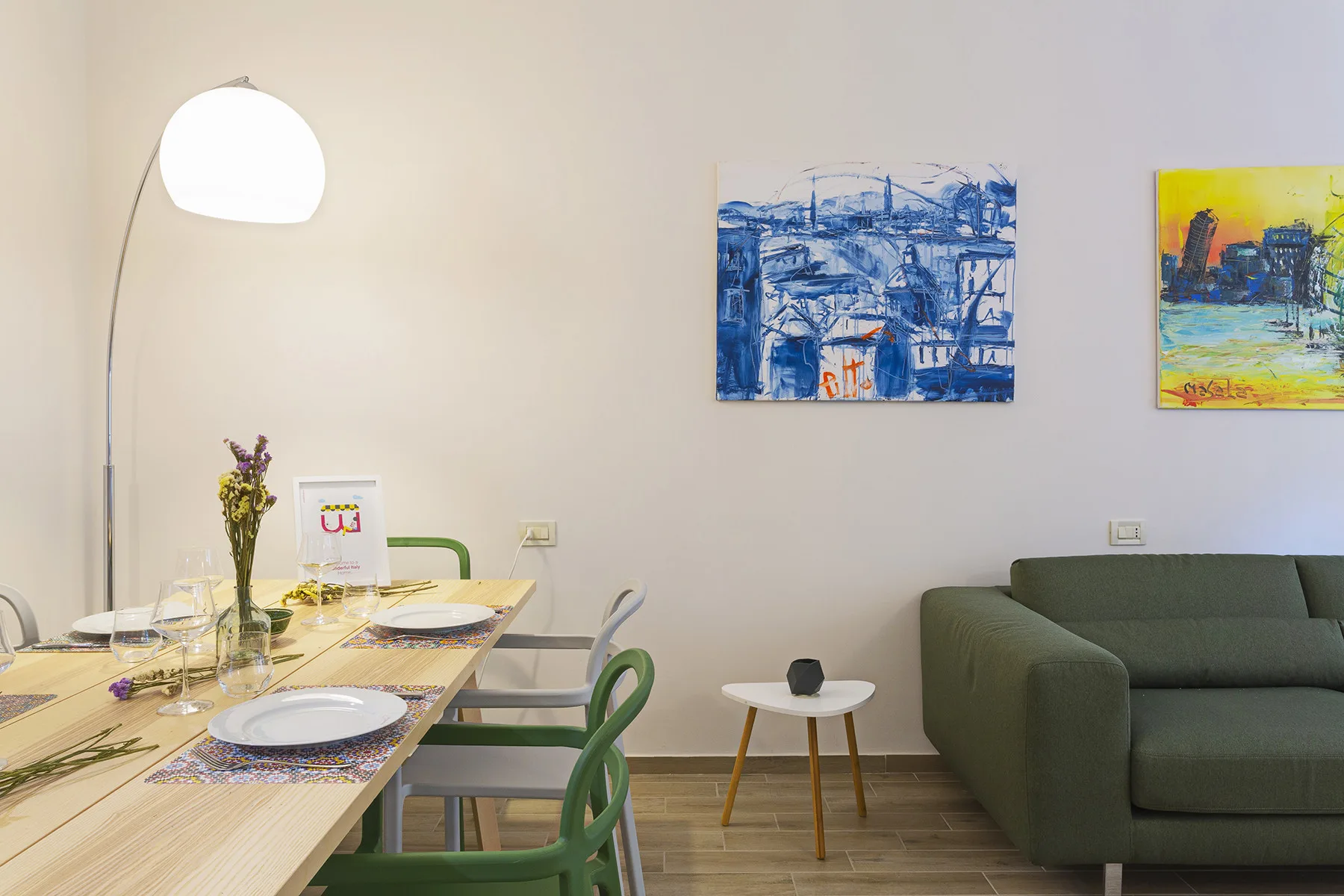 A dining area with a table, seating, and a green sofa beneath colorful wall art.