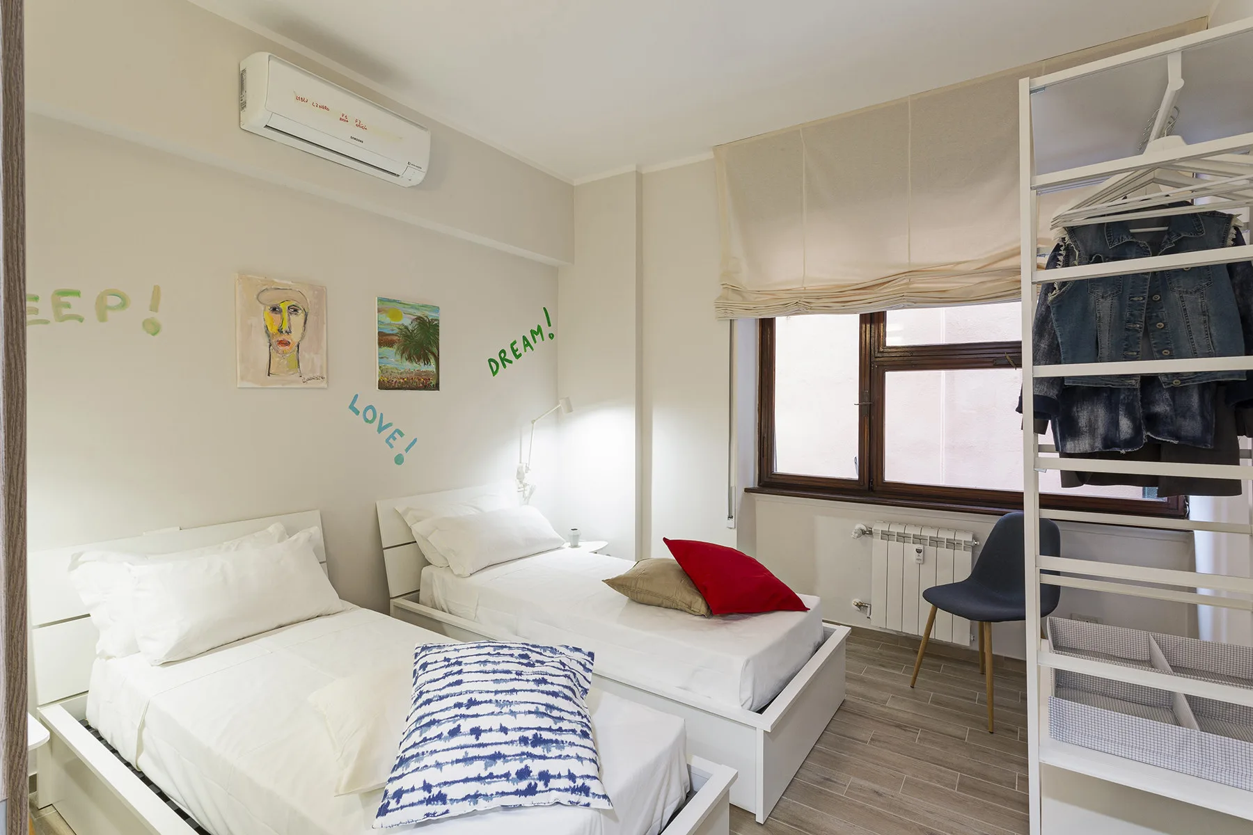 A bright bedroom with two beds, a window, and an open clothing rack.
