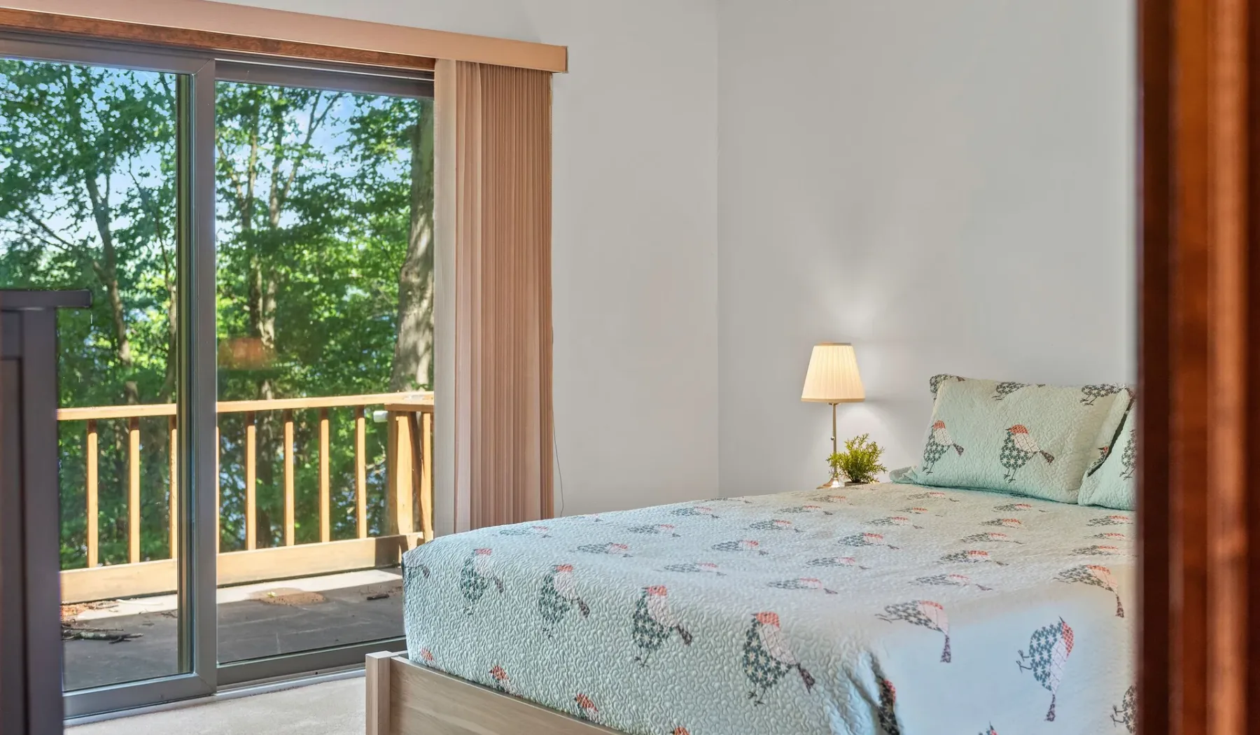 Dormitorio con una cama junto a una puerta corrediza de vidrio que da a árboles frondosos.