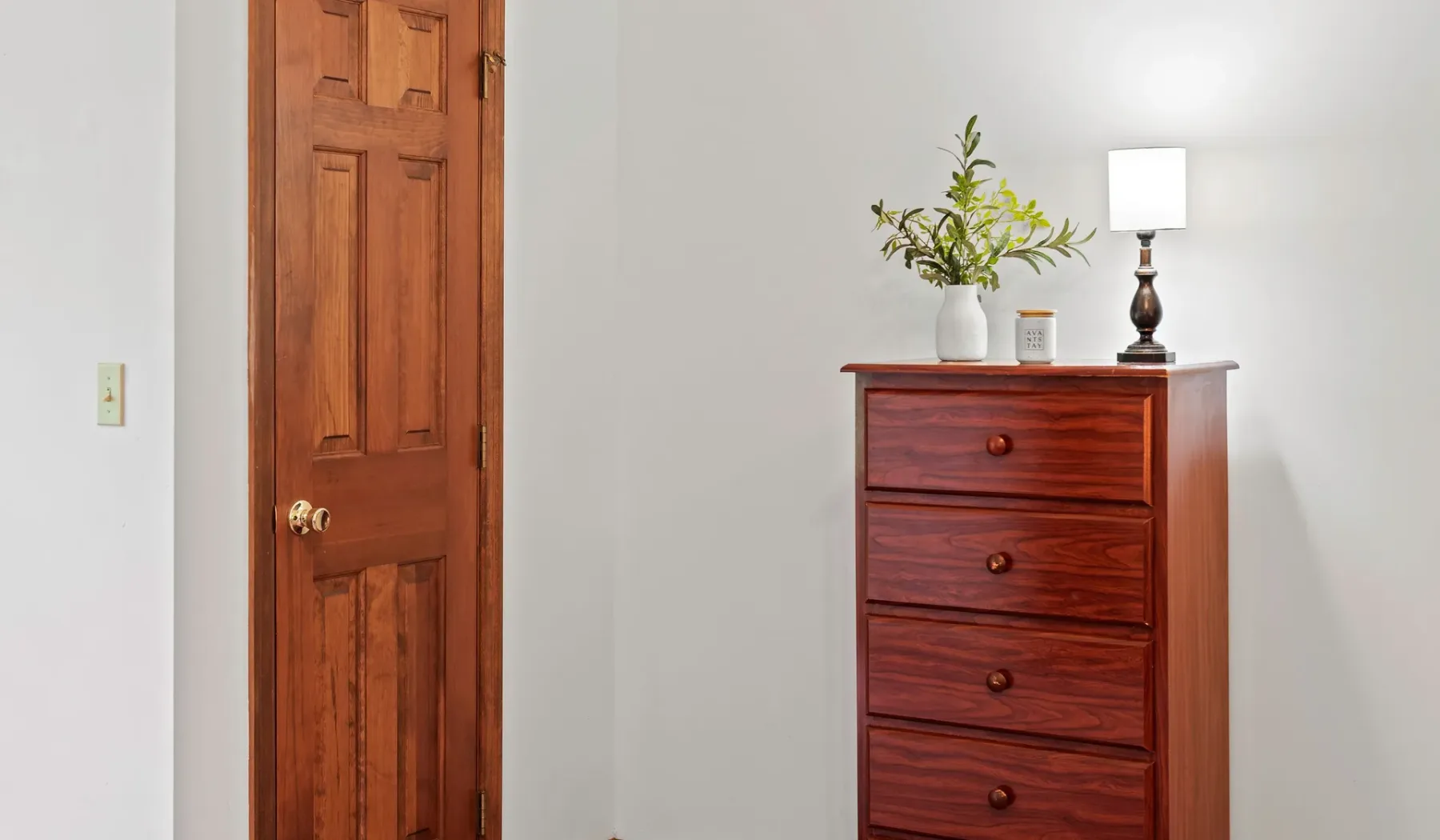 Una puerta de madera está junto a una cómoda con una lámpara y flores encima.