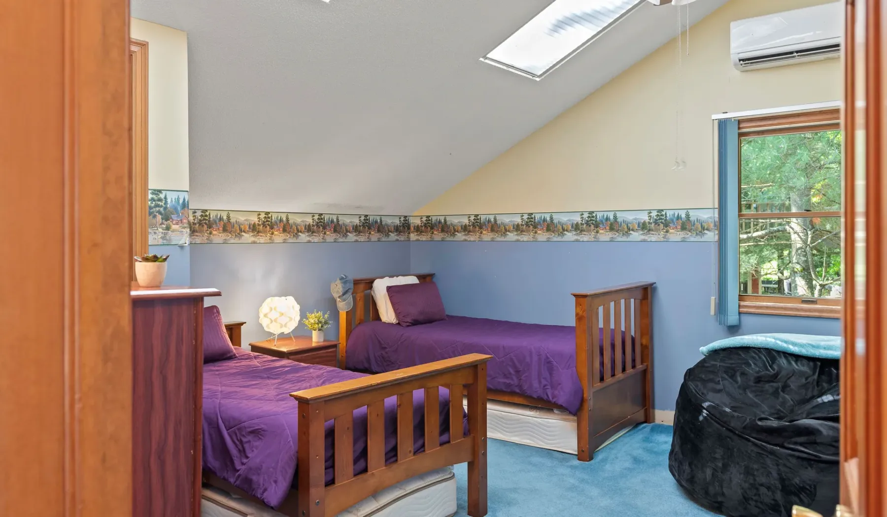 Un dormitorio con ropa de cama morada, camas con marco de madera y una ventana con vista a la vegetación.
