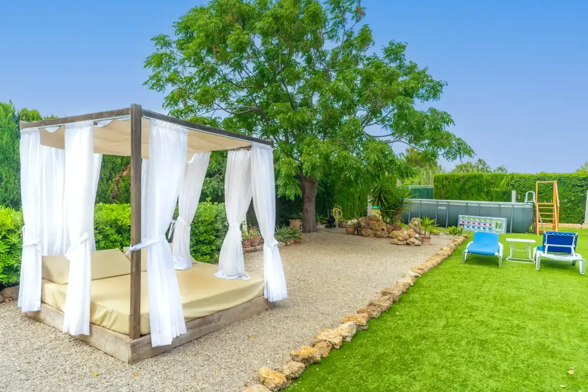 A garden with a curtained outdoor daybed beside a lawn and swimming pool.