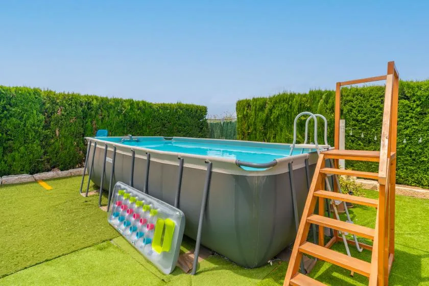 An outdoor above-ground pool sits on artificial grass beside a tall hedge.