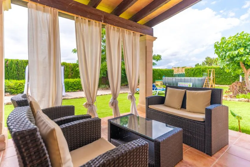 A covered outdoor seating area has wicker furniture, curtains, and a glass-topped table.