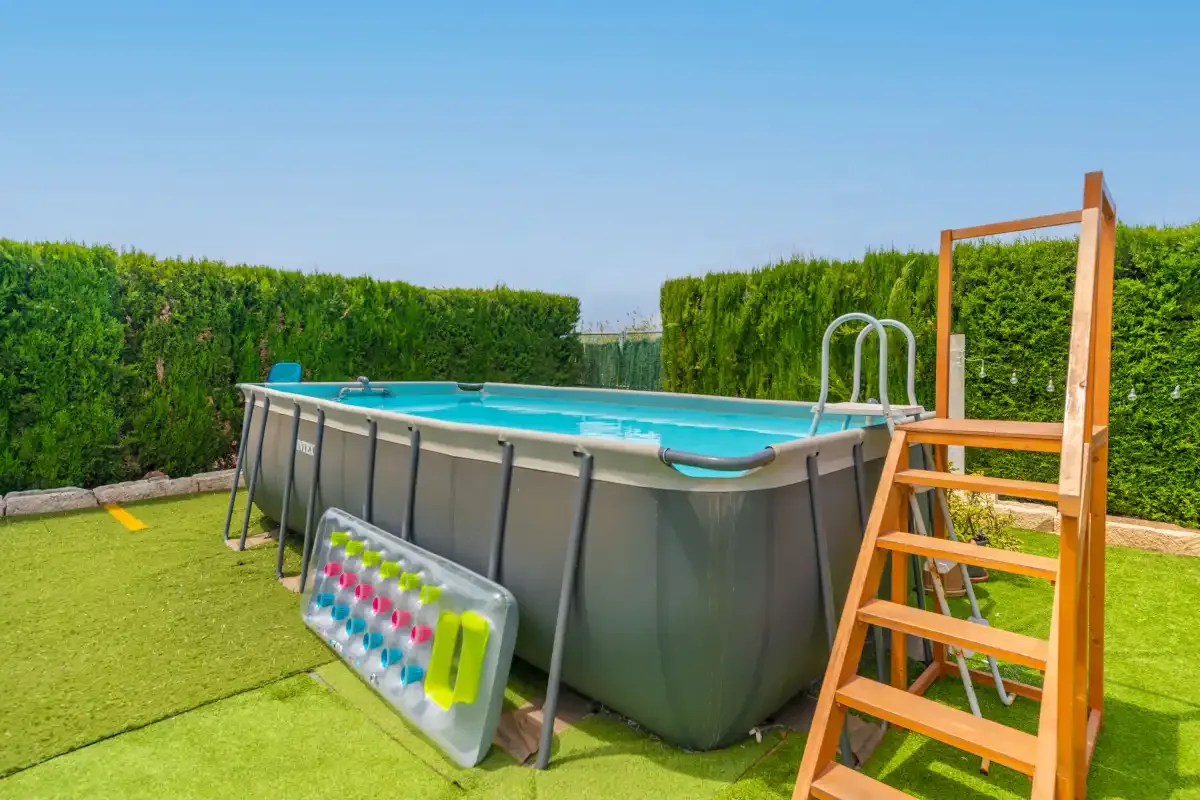An above-ground swimming pool stands on a lawn beside tall hedges.