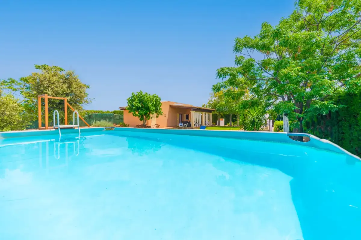 A bright outdoor swimming pool sits beside a low building and leafy trees.
