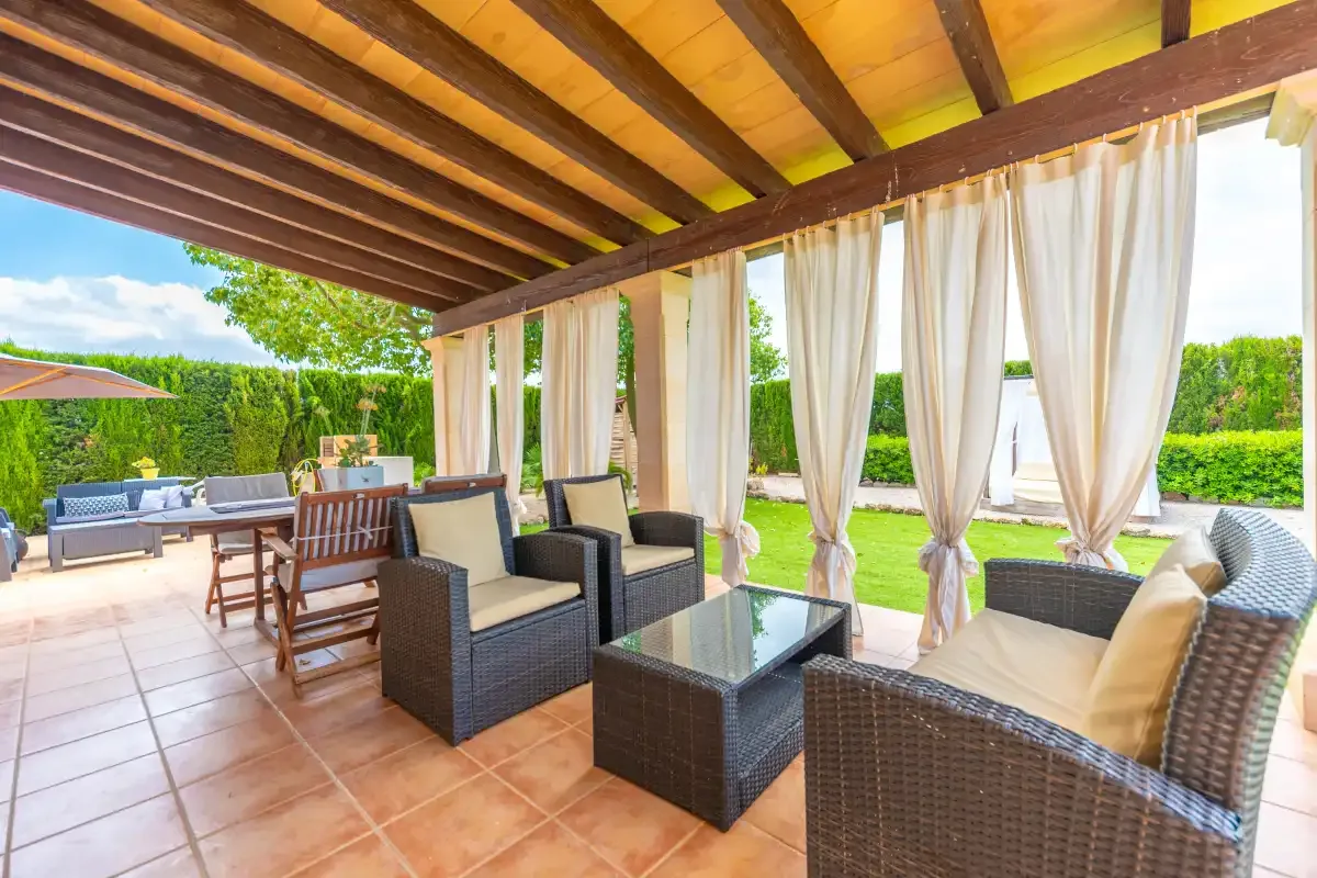 A covered patio with wicker seating and curtains overlooks a green garden.