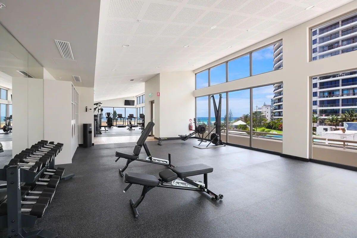 A bright gym with exercise benches and dumbbells beside large windows.