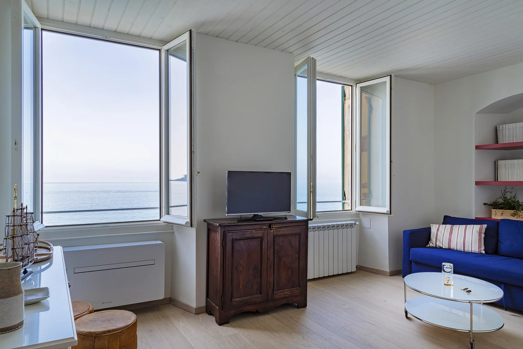 Una sala luminosa con ventanas abiertas frente al mar y una zona de asientos azules.