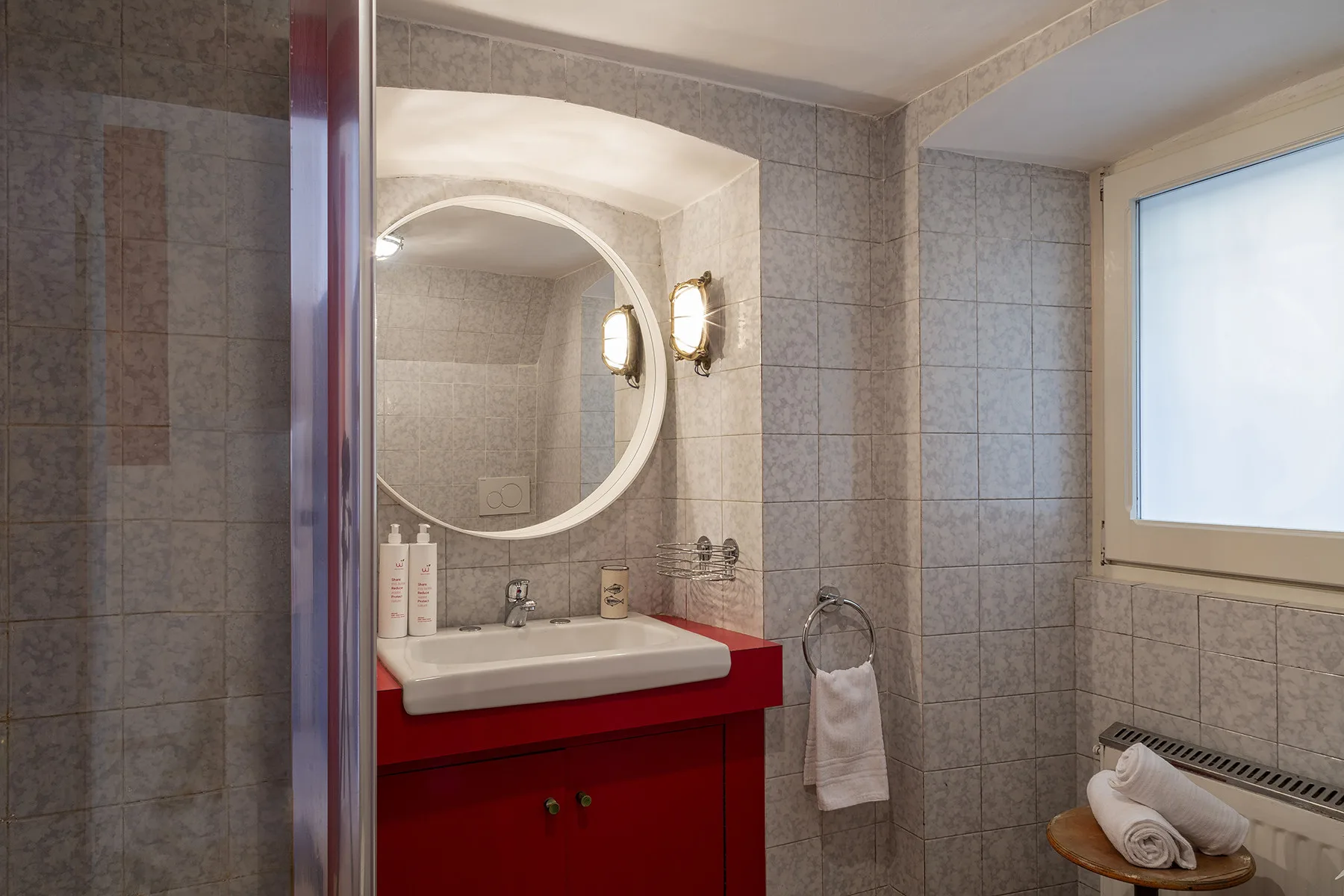 Un baño con azulejos, tocador rojo, espejo redondo y lavabo.